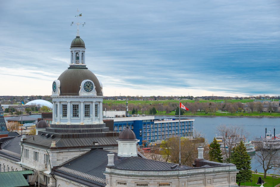 A panoramic view of Kingston riverside showing a historic building with a large domed clock tower and smaller rooftop turrets in the foreground, with construction equipment and scaffolding visible on the rooftop. Behind, there is a modern blue multi-storey building located near the water’s edge, which is a river with boats docked along the bank. The background features a flat landscape with green fields, trees, and a cloudy sky, suggesting a daytime scene. This scene illustrates typical elements involved in house removals and furniture transport, with a focus on the riverfront environment often encountered during home relocation projects, with Kingston companies potentially managing logistics around such area.