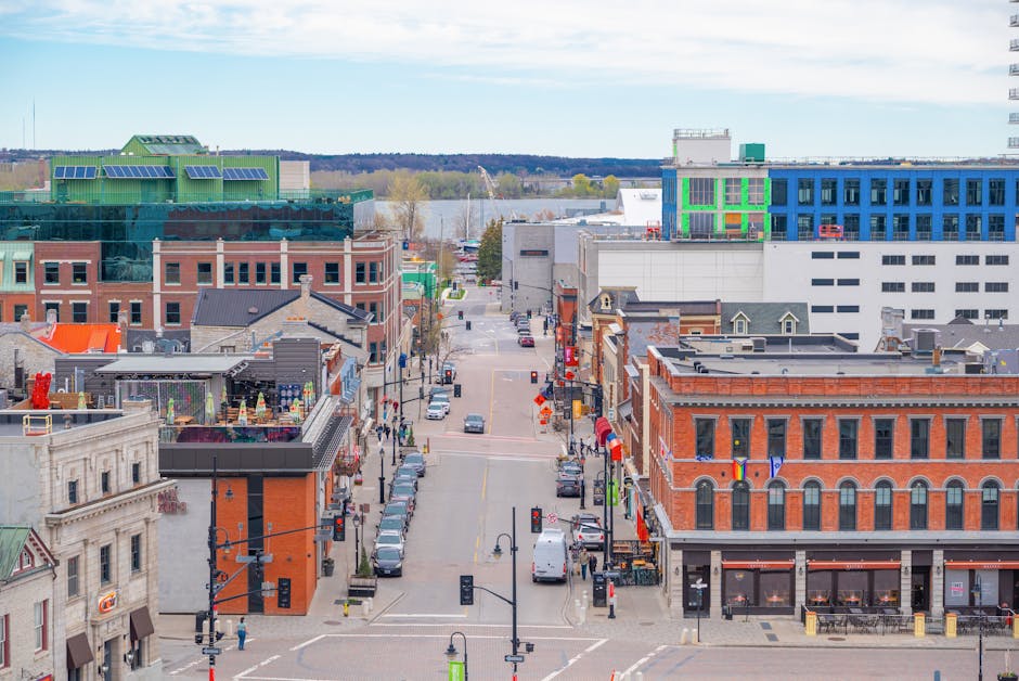 A wide aerial view of a city street scene showing a mix of historic and modern buildings, with several parked cars along the curb and pedestrians walking on the sidewalks. In the foreground, there are large cardboard boxes and wrapped furniture pieces prepared for a home relocation, with some household items visible on a rooftop terrace. The loading process appears to involve a moving van parked nearby, partially visible on the pavement, with a hand truck and moving straps on-site. The scene is illuminated by daylight under partly cloudy skies, illustrating organized furniture transport and packing activities associated with professional removals, as seen in Kingston, as referenced by Removal Companies Kingston. The street extends towards the river in the background, revealing a mix of commercial and residential architecture, emphasizing ongoing moving logistics within an urban environment.