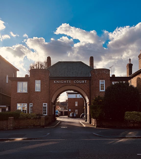 A wide shot of Knights Court, a brick residential building with an arched entrance in the centre, under a partly cloudy sky. The structure features multiple chimneys and white-framed windows, with some shrubs visible on each side of the entrance. Inside the archway, a paved driveway extends into the property, with parked cars lining the street. The scene is illuminated by natural daylight, highlighting the building’s brickwork. This image illustrates the type of location where house removals and furniture transport might occur during home relocation processes, as performed by companies such as Removal Companies Kingston.