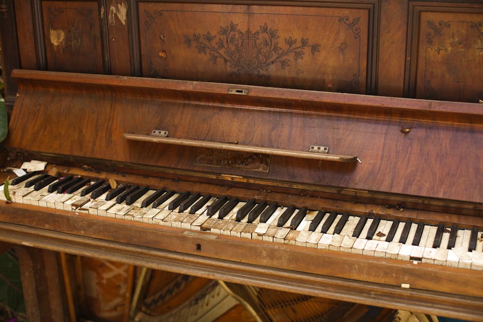 A close-up view of an old, wooden upright piano with an open lid, revealing chipped and stained black and white keys in a state of disrepair. The piano is situated indoors, possibly in a storage area or during a home relocation process. The wood finish is worn, showing scratches and marks, with some decorative floral patterns visible on the panel above the keyboard. Surrounding the piano, there are scattered items including leaves and small debris, indicating it has been moved or stored for a period. The background features a wooden wall or cabinet, contributing to a vintage or antique environment. This image depicts the detailed condition of a piano being prepared for transport or removal, aligning with services offered by Removal Companies Kingston in house removals and furniture transport during a packing and moving process.