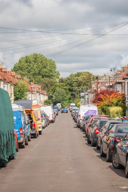 A quiet residential street with parked cars on both sides, including a mix of small hatchbacks, sedans, and larger vans, some of which have cardboard or protective covers on parts of their exterior. The street features typical terraced houses with brick facades, small front gardens, and tiled roofs. In the background, several trees with green foliage line the street, while the sky overhead is overcast with grey clouds. Visible overhead power lines run across the scene, and a few vehicles are in the process of being loaded or unloaded by Removal Companies Kingston as part of a house move, with furniture blankets, straps, and packing materials appearing on some items, indicative of a furniture transport and home relocation process in progress.