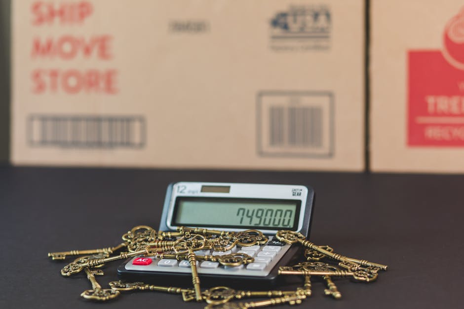 A calculator displaying the number 749800 is placed on a black surface, surrounded by a scattered pile of small, gold-colored keys with intricate designs and bows. Behind the calculator are partially visible cardboard boxes with printed labels and barcodes, suggesting a moving or packing environment. The boxes are positioned on the edge of a doorway or entryway, with the scene illuminated by soft, natural lighting. This image illustrates the process of home relocation, with focus on the packing materials and the logistical aspects of moving, which relate to professional removals services provided by Removal Companies Kingston, especially in the context of furniture transport and loading during house removals.