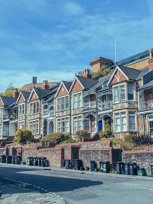 A quiet residential street with parked cars on both sides, including a mix of small hatchbacks, sedans, and larger vans, some of which have cardboard or protective covers on parts of their exterior. The street features typical terraced houses with brick facades, small front gardens, and tiled roofs. In the background, several trees with green foliage line the street, while the sky overhead is overcast with grey clouds. Visible overhead power lines run across the scene, and a few vehicles are in the process of being loaded or unloaded by Removal Companies Kingston as part of a house move, with furniture blankets, straps, and packing materials appearing on some items, indicative of a furniture transport and home relocation process in progress.