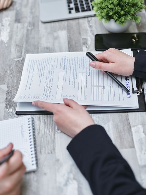A person dressed in a dark suit is holding a pen and reviewing a document labeled 'Insurance Policy' on a clipboard, with their other hand pointing at the paper. The scene is set on a wooden work surface that appears to be in an office or staging area for house removals. Nearby items include a small notepad, a black smartphone, and an open laptop, all arranged on the table. To the right, there is a potted green plant with dense foliage, and an orange and white box, possibly for packing supplies, can be seen in the background. The person's hands are positioned over the document, indicating active review or signing related to moving insurance or compliance. The setting suggests a professional environment where attention to detail and proper documentation are essential, aligning with the services provided by Removal Companies Kingston involved in home relocation and furniture transport. The lighting is natural and even, illuminating the workspace clearly.