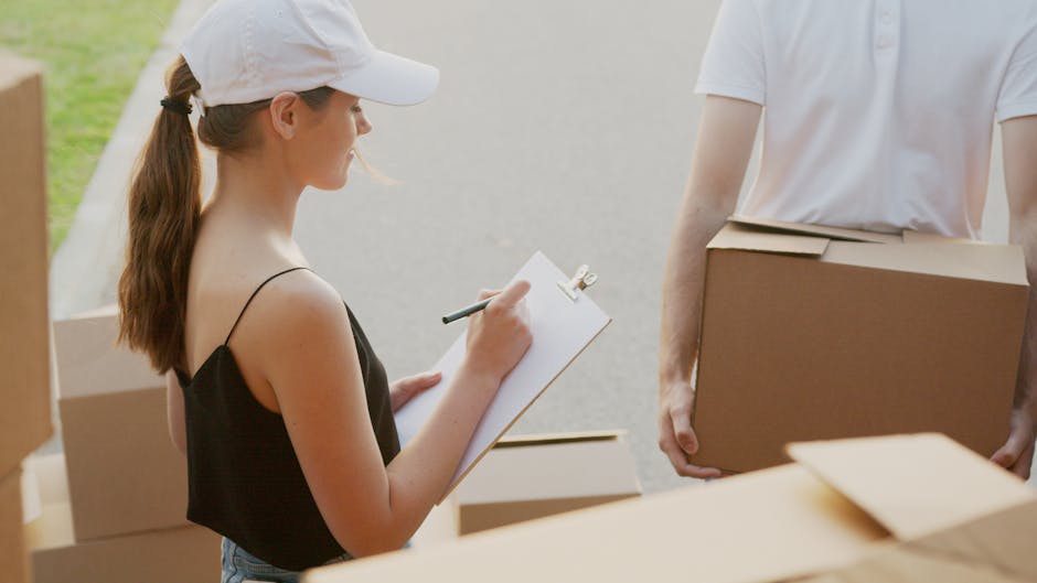 A young woman wearing a white cap and a sleeveless black top is inside a property, standing on a paved area near open cardboard boxes filled with packing materials. She is holding a clipboard and pen, taking notes as part of the home relocation process managed by Removal Companies Kingston. Next to her, a person in a white T-shirt is partially visible, carrying a large sealed cardboard box. Surrounding them, there are more packed boxes, some stacked and others on the ground, indicating an organized packing and moving operation. The scene is well-lit, likely during daytime, and shows the indoor-outdoor transition typical of a household moving setting. The background includes a blurred view of the outdoor environment, emphasizing the context of furniture transport and loading for a house removal or local moving service, aligning with the content related to insurance and compliance for house removals in Kingston.