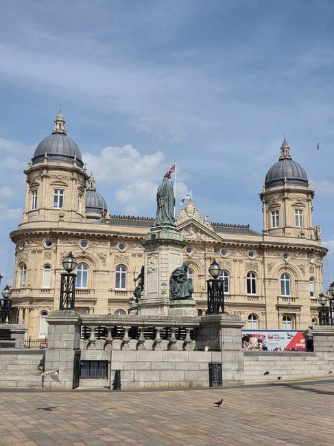 A historic stone building with elaborate architectural features, including four domed towers, is visible in the background of a street scene. In the foreground, there is a stone monument with a bronze statue of a seated figure, surrounded by black wrought-iron lamp posts and a low stone wall with decorative balustrades. The pavement is made of large square tiles, and a single bird is walking on the street. The scene appears under a partly cloudy sky, with a few birds flying overhead. Although the image does not depict moving or packing items directly, it is associated with the context of house removals and city logistics, such as planning for large vehicle access or permits through Kingston Council, as mentioned on the webpage of Removal Companies Kingston.