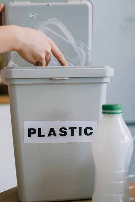 A close-up image of a grey plastic waste bin labeled 'PLASTIC' in bold black letters, positioned indoors next to a white plastic bottle with a green cap. The bin's lid is open, revealing a clear plastic container inside. The scene suggests the sorting and disposal of recyclable materials as part of a home relocation or packing process, with the bin placed on a wooden surface. The background is blurred, indicating a typical domestic environment, and the image is related to removals and waste management services offered by Removal Companies Kingston, especially following Kingston Council waste rules after furniture and household goods are moved.