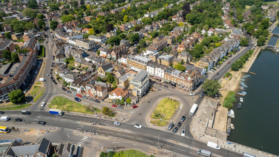 An aerial view of a seaside town showing a busy street with several vehicles, including cars, vans, and trucks, parked or driving along the road. Adjacent to the street, there is a small roundabout with a patch of grass and surrounding parking areas. The town features a mixture of residential and commercial buildings with brick facades, some with storefronts facing the street. On the right side of the image, a waterfront promenade with a pathway and moored boats is visible alongside the water’s edge. Trees and greenery are scattered throughout the town, creating a pleasant urban landscape. The scene captures the active environment typical of a home relocation and furniture transport operation, with some vehicles and equipment possibly related to removals services by Removal Companies Kingston, supporting efficient packing, loading, and transit of household items near Kingston Bridge in Kingston KT1.