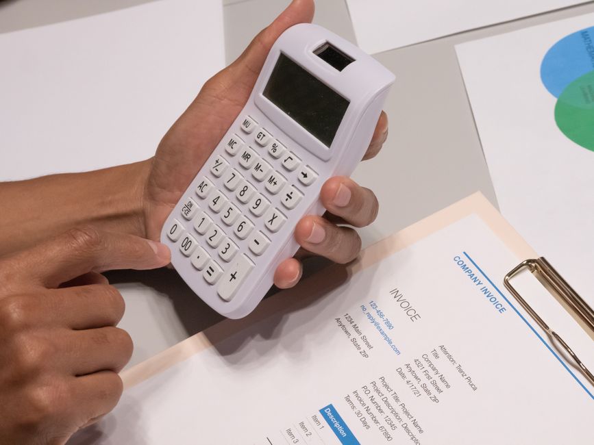 A flat lay image shows a person’s hands on a light blue surface, with one hand holding a pen and the other reaching towards a calculator displaying a numerical total. Surrounding the hands are three
