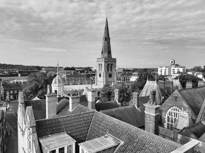 Black and white photograph of a cityscape featuring a large church with a tall, pointed spire and a clock face, surrounded by various rooftops covered in tiles. Some rooftops have chimneys and small dormer windows. In the foreground, there are several building rooftops, some with visible chimneys and flat sections, with trees and additional buildings extending into the background. The sky above is partly cloudy, with a mix of light and darker clouds. The image captures an urban area during daytime, highlighting historic and modern architecture, and provides a clear view suitable for illustrating house and building removals or relocation processes, as reflected in the context of [PAGE_TITLE] linked to [COMPANY_NAME]’s removals services.