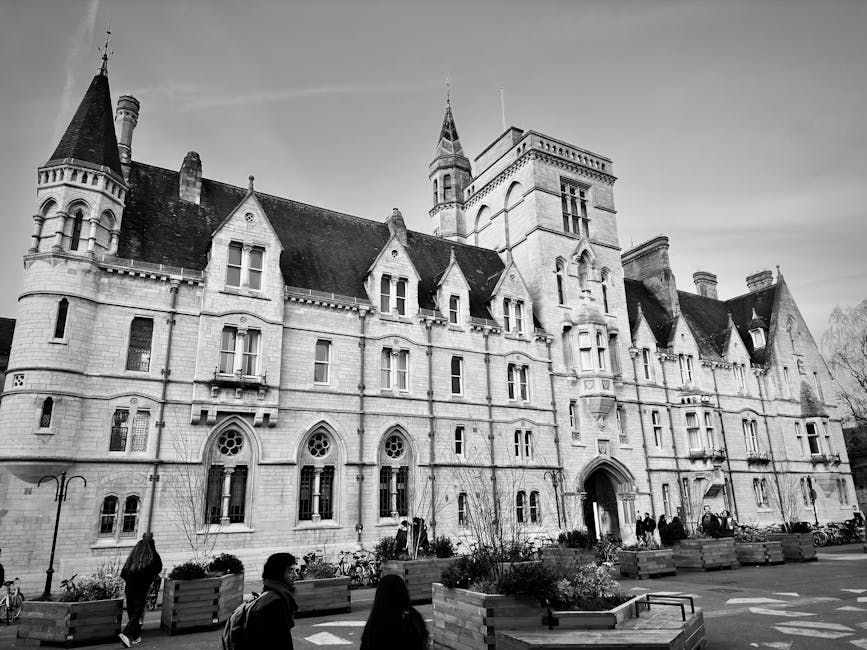 Black and white photograph of a large historic building with multiple turrets, gabled roofs, and tall, narrow windows, situated on a paved area with planters containing small trees and flowers. Several people are walking in front of the building, and bicycles are parked along the pavement. The scene captures an exterior view of the building during daylight, with a clear sky overhead. The image relates to house and building removals, illustrating the type of architecture involved in the relocation process. The presence of pedestrians and bicycles indicates a busy environment where transportation of furniture and belongings might be handled by professional services such as Removal Companies Kingston, especially when planning student relocations to nearby Kingston University halls, as referenced in the webpage 'KT2 student removals to Kingston University halls, KINGSTON'.