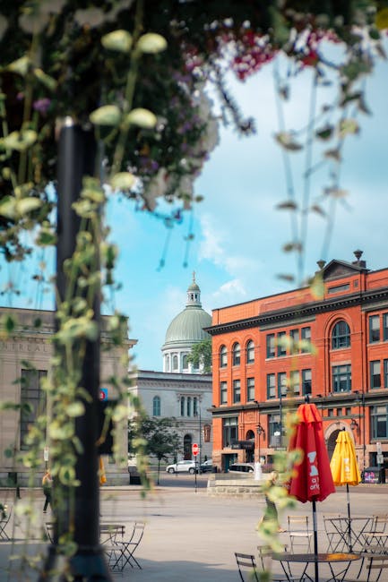 The image shows a city square with a historic dome and a red-brick building in the background under a partly cloudy sky. In the foreground, there are blurred green foliage and hanging flower baskets, framing the scene. On the pavement, outdoor tables and chairs are visible, along with closed red and yellow umbrellas. The area appears quiet with minimal pedestrian activity. Although this image does not depict house removals or moving services directly, it provides contextual urban scenery that could relate to relocation or transport logistics in a city setting, aligning with the services offered by Removal Companies Kingston at Surbiton station for same-day house moves within Kingston.