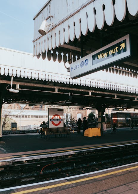 The image shows a railway station platform under a large, white, scalloped canopy with a black underside, providing cover for waiting passengers. In the foreground, train tracks are visible, with yellow safety lines marking the edge of the platform. Several people are standing and sitting on wooden benches, some near luggage or bags, indicating a busy scene typical of home relocation activities involving train travel. A prominent sign with a green background and white text points to the exit and lift, mounted on the canopy structure. The station environment features modern amenities, with an urban backdrop suggesting proximity to residential or commercial areas. As part of the moving process, Removal Companies Kingston could utilize such platforms for efficient furniture and packing material transport between locations during a house removal or furniture transport service, ensuring safety and punctuality for same-day moves at Surbiton station.
