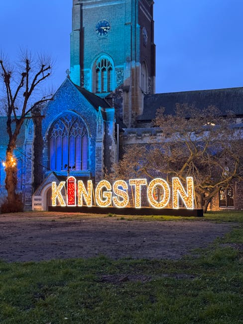 The image shows a large, illuminated sign spelling out 'KINGSTON' in bold, red and white lit-up letters positioned on the grass in front of a historic stone church with tall, pointed arched windows, illuminated in blue lighting. The church is situated in a town square with leafless trees nearby, indicating a winter setting during the evening or night. In the foreground, there is an uneven dirt and grass surface, suggesting ongoing home relocation or furniture transport activities associated with house removals. The scene is part of a professional removals service in Kingston, with the church serving as a recognizable local landmark. The lighting highlights the architectural details of the church and the sign, subtly supporting the theme of moving and packing services provided by Removal Companies Kingston.